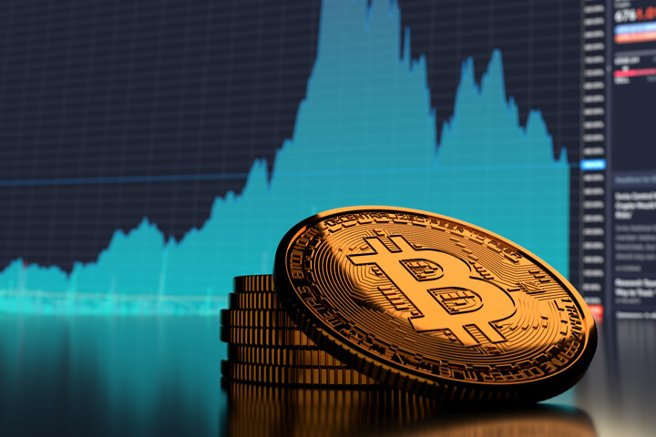 Small stack of Bitcoins in front of blue market area chart showing sharp rise and fall in Bitcoin price