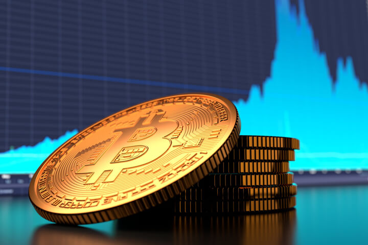 Small stack of Bitcoins in front of monitor showing blue area chart with cryptocurrency price fluctuations