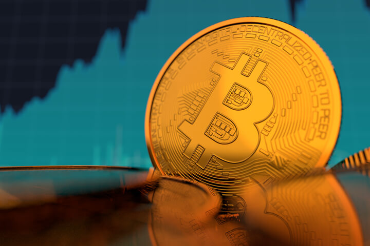 Single Bitcoin on edge in front of monitor showing increasing stock price of Bitcoin and BTCUSD