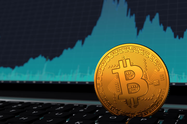 Bitcoin vertical on laptop keyboard in front of blue price volatility area chart on screen