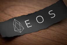 EOS altcoin cryptocurrency logo printed on torn piece of black scrap paper lying on woodgrain surface