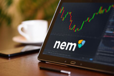 Tablet on desk with coffee cup and cell phone showing Nem cryptocurrency logo and stock price candlestick chart