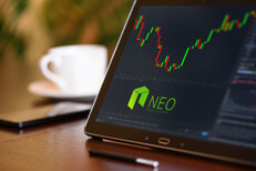 Tablet on desk with coffee cup and cell phone showing Neo cryptocurrency logo and stock price candlestick chart