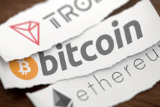 Tron, Bitcoin, and Ethereum cryptocurrency logos printed on torn pieces of white scrap paper on woodgrain surface