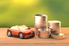 Small red toy car next to stacks of coins on wood table with nature background