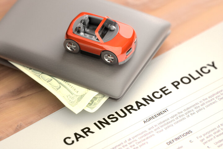 Toy car sitting on top of wallet with car insurance policy papers on wood table