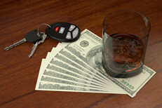 Glass of whiskey and car keys on top of fanned one hundred dollar bills representing the cost of a DUI conviction