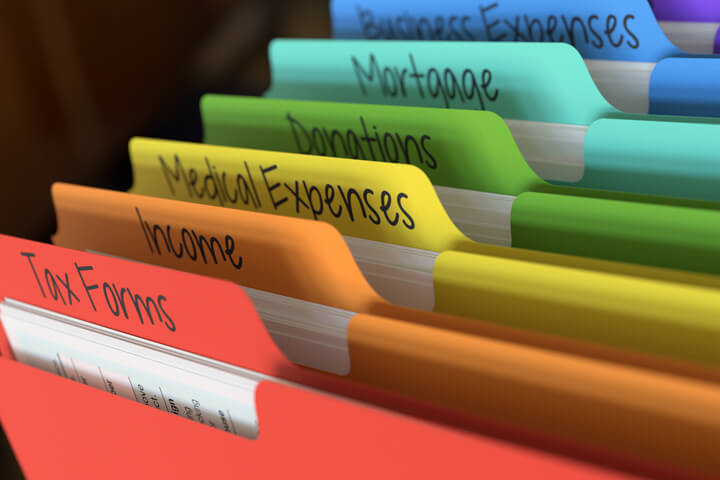Rainbow-colored tax folders in filing cabinet with tab text for income, expenses, deductions, and mortgage