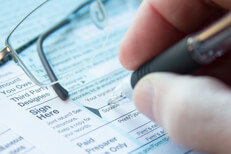 Photo of hand holding pen signing tax form with reading glasses