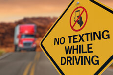 Yellow road sign reading No Texting While Driving with no texting icon and oncoming semi truck in background